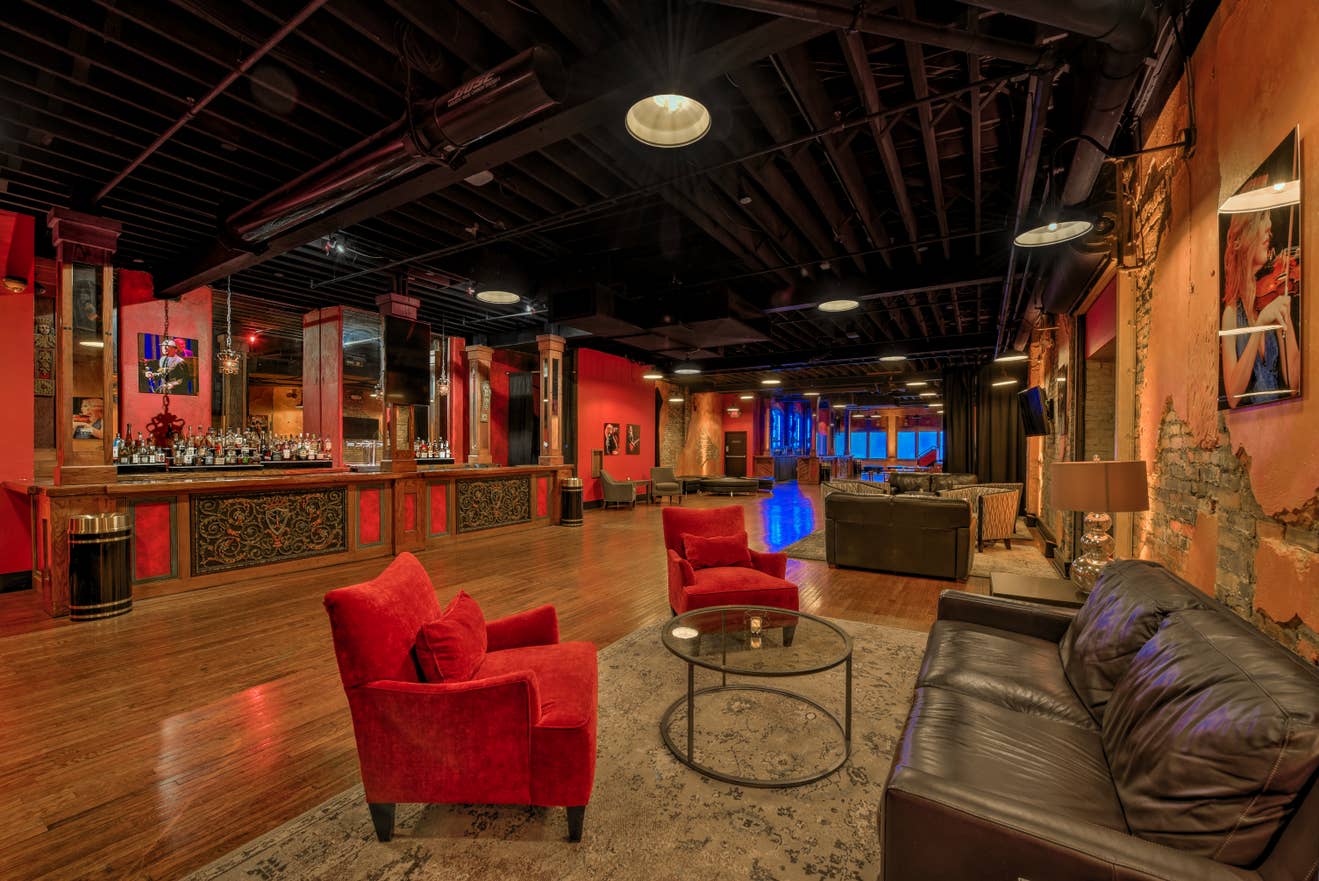 The Marquee Lounge at the Louisville Palace, featuring plush chairs, warm lighting, exposed brick and beams, a full-service bar, and bold red patterned carpet for a stylish yet relaxed vibe.